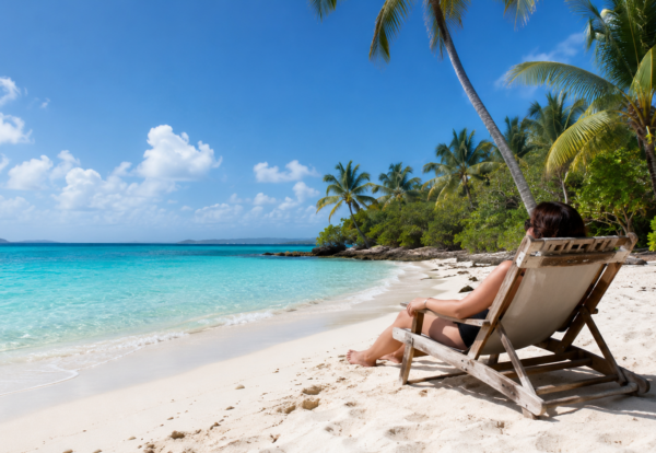 Person relaxing in a beach chair on a white-sand Caribbean beach overlooking clear turquoise water.
