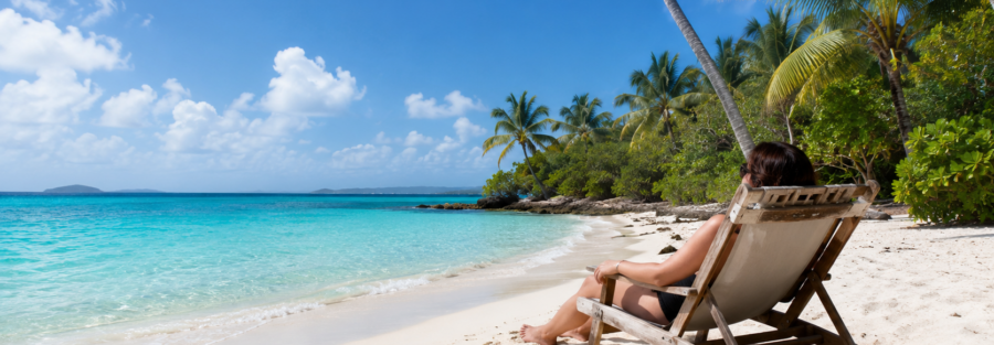 Person relaxing in a beach chair on a white-sand Caribbean beach overlooking clear turquoise water.